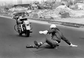 Black-and-white photograph showing a motorcylist wearing gloves and helmet, lying on the road, raising themselves up with their hand, with the motorcycle stood in the road nearby.