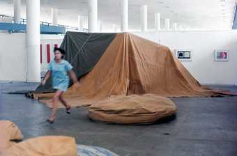 In the white columned exhibition space in front of wall-mounted images is an amorphous installation made from brown and black material - a boxy tent-like shape and another beanbag/pancake shape nearby. A person moves across the space, blurry in motion.