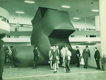 Visitors and security guards stand next to one of a series of enormous inflatable sculptures, this one forming an 'X' nearly reaching the full height of the exhibition space.