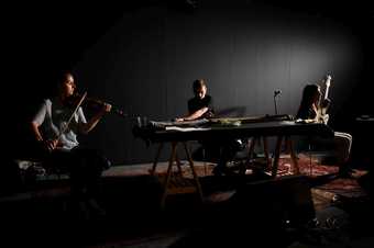 Three musicians play a violin, a long guitar-like instrument and a bass guitar in a darkened room
