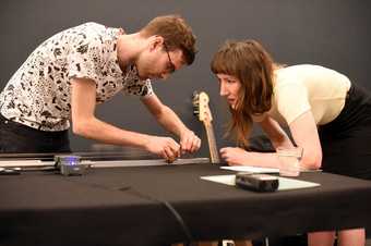 Two musicians talk while one of them tunes the strings of a guitar-like instrument resting on a table