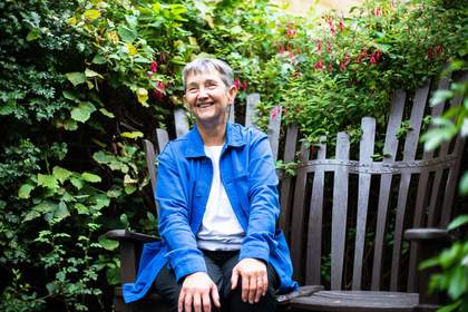 woman sitting on a metal bench, smiling wearing a blue shirt and there is foliage all around the bench.
