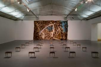 A photograph of a fabric wall hanging on the back wall of a gallery with 5 rows of books perched on top of individual stands across the room.