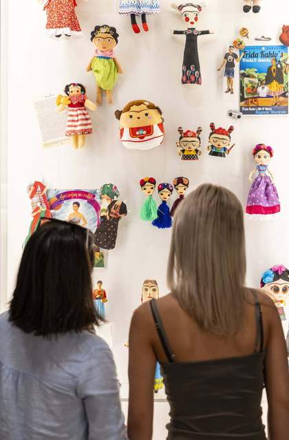 a person with brown hair next to a person with blonde hair in front of many different types of dolls of Frida Kahlo
