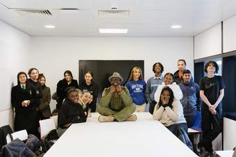 A photograph of young people stood around a table with a man sat cross legged in the centre