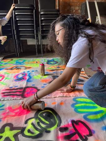 A photograph of a young person spray painting on a piece of fabric on the floor.