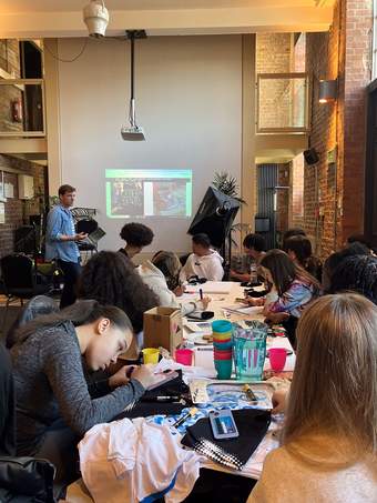 A group of young people drawing and doing craft activities at a table whilst someone is speaking in the background in front of a projected screen.