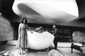 Image of a woman in front of a large sculpture that is in two parts, one part raised up to the ceiling.