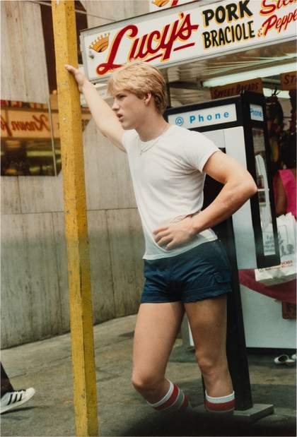 A man leans on a post on a busy New York street. He has a hand on his hip. He wears a white t shirt, short blue shorts and calf height white and red socks.