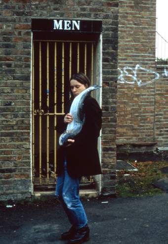 Photograph of Sarah Lucas stood in front of the gate of a public men's lavatory holding a salmon fish against her body as if slung over her left shoulder.