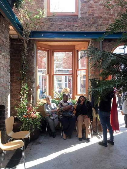 A photograph of three people sat on chairs in front of a window looking up and talking to a person who is standing. There are two empty chairs on the left and exposed brickwork with blue steel beams above.