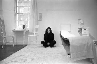 Black and white image of a person with black hair and black clothes sitting in a room with halved white furniture
