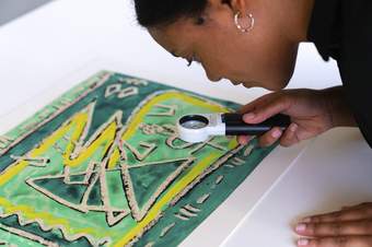A person with dark skin looks at a green and yellow abstract artwork with a magnifying tool