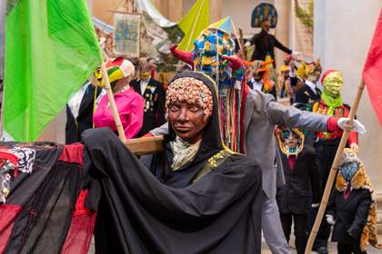 A woman with a headdress and a cloak carrying a pole over her shoulder.