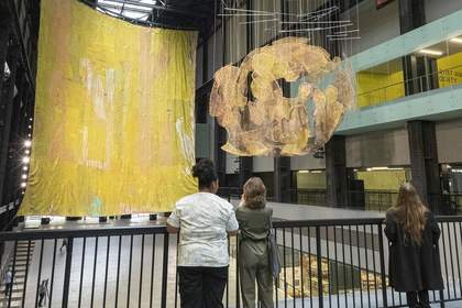 Three people standing on a bridge at Tate Modern looking at a large gold installation