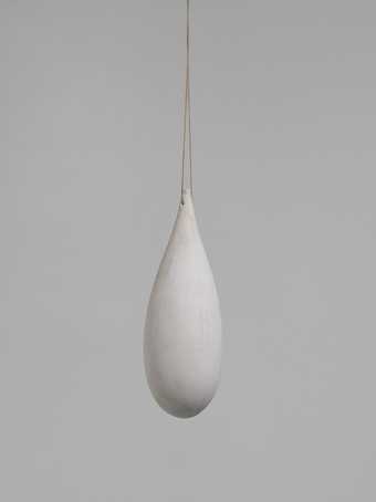 A drop-like sculpture hanging from a thin support against white backdrop