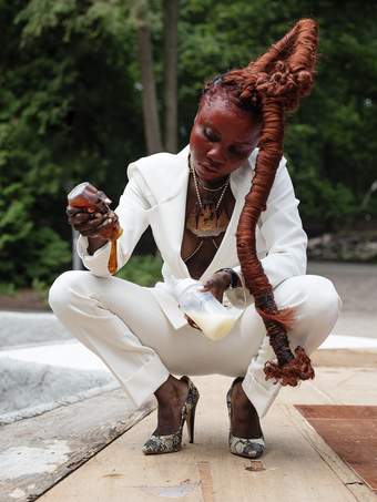 A photograph of a woman in a white suit and snake-print heels squatting down whilst holding a bottle of milk and a tub of syrup.