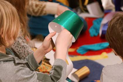 A child holding a strip of curled shiny green paper, against a background of other people making with brightly coloured materials sitting on mats.