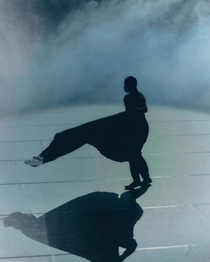 A portrait oriented photograph of a performer on a smoke filled stage. The performer is silhouetted in bright white lighting and waves a piece of fabric.
