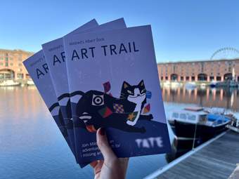 A person holding three booklets with the title 'Mildred's Albert Dock Art Trail' in front of the Royal Albert Dock in Liverpool. There is water in the background with boats and buildings under a clear blue sky.