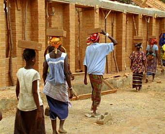 A still from a film showing people walking behind each other carrying bricks on their head.