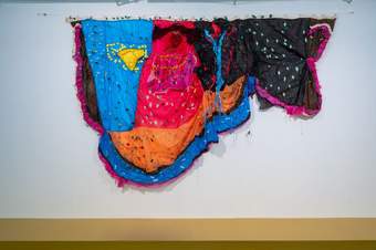 A photograph of a fabric installation hanging on a wall. The artwork is made with blue, yellow, red, pink and orange coloured material.