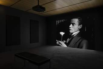 View of gallery with a projection of a black and white film in front of a bench, showing a man holding a flower.