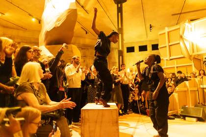 Photograph of nora chipaumire singing into a microphone, whilst dancer Yinka Esi Graves stands atop a wooden box.  They are surrounded by a large audience watching.