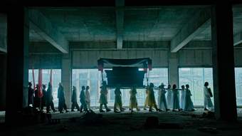A procession of people carry a coffin through a dark industrial building