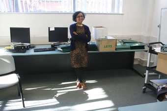 A person standing in an office space, with arms crossed and smiling, with computers, paper files and cardboard boxes on the desk behind them.