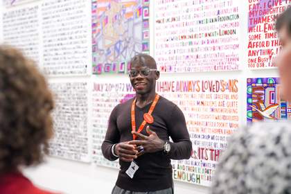 A man wearing a black top smiling and gesticulating, with Tate lanyard and the blurred image of two people looking at him in the foreground.