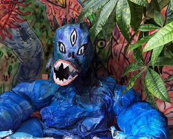 An image of a blue monster sat at a table with leaves in the background