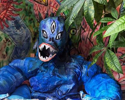An image of a blue monster sat at a table with leaves in the background
