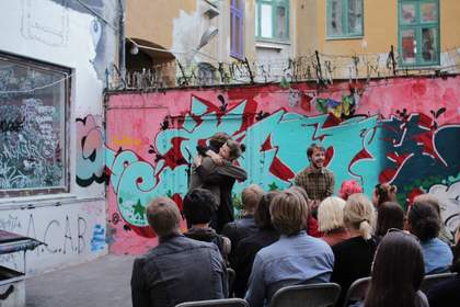 A photograph of two people hugging whilst another person addresses an audience. There are the backs of the audiences' heads in the foreground.