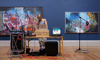 Joshua Woolford performing at Tate Britain, standing in front of their laptop and surrounded by TV screen, a microphone and and cables.