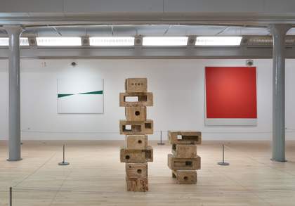 In the foreground is a sculpture of bricks stacked up on top of each other. In the background on the left is a green and white abstract work and on the right a bright red square painting.
