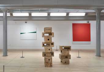 In the foreground is a sculpture of bricks stacked up on top of each other. In the background on the left is a green and white abstract work and on the right a bright red square painting.