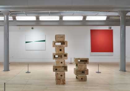In the foreground is a sculpture of bricks stacked up on top of each other. In the background on the left is a green and white abstract work and on the right a bright red square painting.