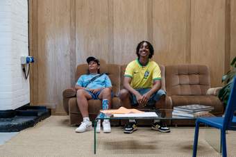 Two young people are sat on a sofa during a photoshoot smiling.