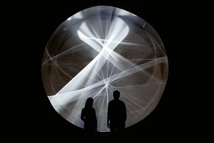 A black rectangle has a circle cut out of the middle, within which we see waves of light. Two silhouettes of people stand in front of it, looking at the work