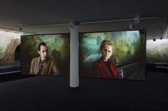 Two large screens showing a film in a room with a large mural painting on the walls