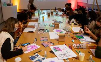 A group of people at a workshop, seated around a long table, drawing using a variety of materials.