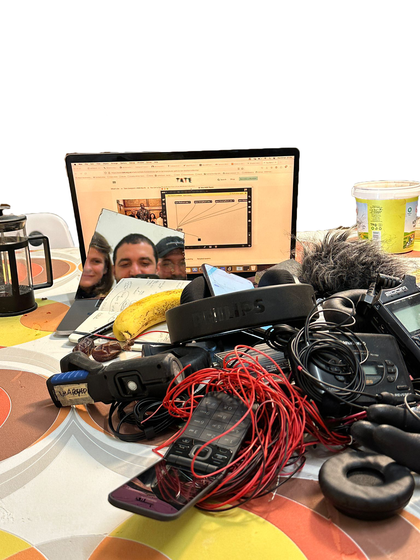 A table with headphones, microphone, recorders, a flip phone and cables as well as a french press coffee pot, open notebook and a banana. Behind these items there is a mirror showing 3 people, one with long hair, one with short hair and one wearing a cap