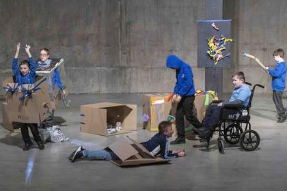 Young people wear costumes built out of cardboard boxes in the tanks. One has a box around his waist as he lies on the ground, looking up. One student in a wheelchair looks at the costumes.