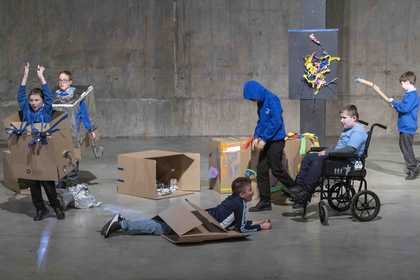 Young people wear costumes built out of cardboard boxes in the tanks. One has a box around his waist as he lies on the ground, looking up. One student in a wheelchair looks at the costumes.
