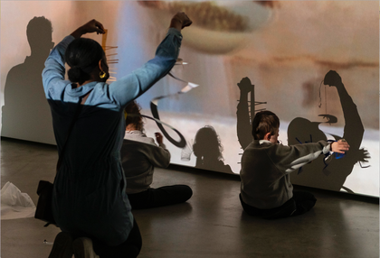 An artist holds mobiles up to a projected video. Two young people sit on the floor, making shadows in the video projection