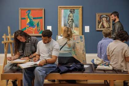 People sit on a bench in a gallery drawing with clipboards, paper and pencils