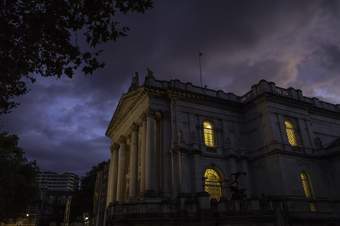 A photo of Tate Britain's millbank entrance from the outside at a diagonal angle view. It is night time and you can the dark eery sky above and trees in the left top corner.