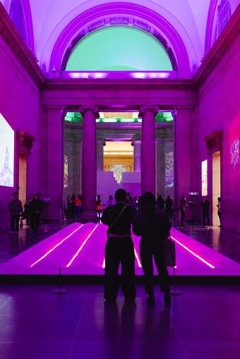 The silhouette of 2 people standing in front of Vong Phaophanit's Neon Rice Fields installation with the room bathed in purple light