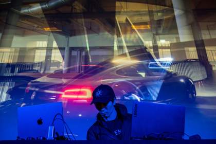 Artist Lawrence Lek standing behind 2 laptops and in front of a projection with layered images of sports cars